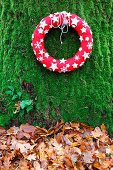 Decorative red wreath with white stars on mossy tree trunk above autumn leaves