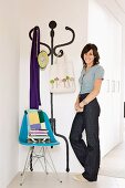 Woman standing next to turquoise shell chair with metal frame in front of coat rack painted on wall, bag on hook and white fitted wardrobes in background