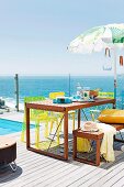 Wooden table, matching bench and yellow chairs on sunny wooden terrace beside pool with sea view through glass balustrade