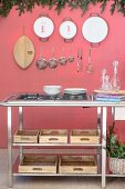 Gas hob on metal-framed counter above wooden boxes on shelves in front of kitchen utensils hung on pink wall