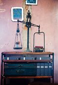 Antique scales on simple, half-height shelving with metal trunk and blue box files against pink wall with damaged plaster