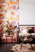 Romantisches Arrangement - Rattan Schemel auf rundem Sisalteppich vor Sitzbank mit Polstern, daneben Blumenstrauss auf antiken, rosa Koffer auf Boden, vor geblümtem Wandbehang