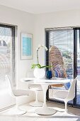 Orchid in spherical vase on Tulip table with matching white shell chairs in corner in front of balcony doors with louver blinds