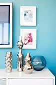 Mirrored vases of various sizes on white sideboard below framed pictures on wall painted pale blue
