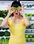 Mixed race woman covering eyes with kiwi slices near refrigerator