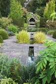 Stone flagged area with narrow water channel, ornamental grasses and pool and traditional pavilion in background in English garden