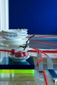 Oriental table setting: cutlery wrapped with red woollen yarn, fruit bowl on plexiglass blocks and stacked plates and dishes in background