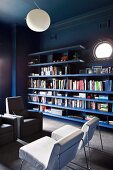 Dark blue bookshelves on dark wall with porthole window contrasting with white easy chairs and white spherical ceiling light