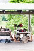 Rustic wooden table and chairs, dog lying on animal-skin rug and wicker sofa with cushions on roofed wooden terrace in summery landscape
