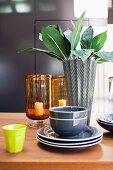 Graues Geschirr mit schwarz-weißer Designer-Vase, zitronengelbem Becher und Glaswindlichtern auf Holztisch arrangiert