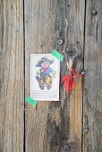 Nostalgische Grafik mit kleiner Cowboyfigur fixiert mit Masking tape und zartem Weinrebenblatt dekoriert an verwitterter Bretterwand