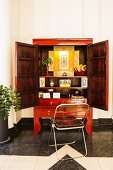 Folding chair in front of red, Chinese wedding cabinet with wide open doors and view of shrine-style arrangement inside