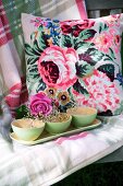 Grüne Schächen mit Schleierkraut und Rosenblüte auf einem Tablett vor Kissen mit Blumenmuster und karierte Decke
