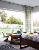 Corner of room with glass walls, partially visible bed and classic, green children's chairs around model boat on play table