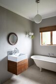 Modernes Waschbecken mit Holzunterbau und minimalistischer Spritzschutz aus Mosaikfliesen unter rundem Spiegel an hellgrau getönter Wand, an Fenster freistehende Badewanne