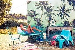 Decoration for outdoor party with combination of azure blue seating, rug and floor cushions in front of palm tree pattern on screen; young woman posing on bench