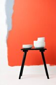 Stacks of white china crockery on black stool against white wall painted with fields of red and grey