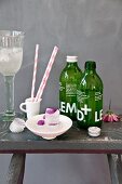 Eiswürfel mit eingefrorenen Cosmea-Blütenblättern, Zitronenlimonade in grünen Flaschen und gestreifte Papierstrohhalme auf rustikalem Schemel