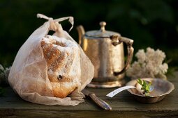 Bundt cake wrapped in paper next to vintage cutlery and teapot on wooden surface