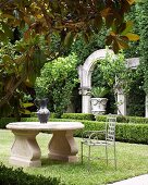 Metal chair at classic stone table in garden; antique columns, arches and urns behind low box hedge