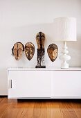 African masks on transparent plastic stand next to white table lamp on designer sideboard