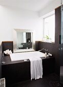 Bathtub in niche built into niche with window, silver-framed mirror leaning against wall, black mosaic tiled on bath surround and walls