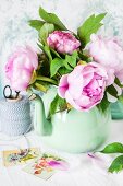 A bunch of pink peonies in a green metal jug against a wallpapered wall