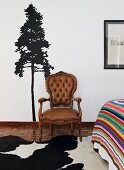 Antique chair with brown leather cover in front of stencilled tree motif on wall, cowhide rug on mosaic wooden floor next to partially visible bed