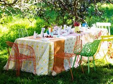 Table set with various tablecloths and colourful metal chairs on lawn in garden