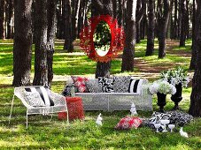 Spiegel mit rotem Metallrahmen an Baum aufgehängt & weiße Outdoor-Möbel aus Maschendraht mit Dekokissen auf einer Waldlichtung