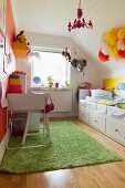 Schreibtisch und Stuhl in Weiß auf grünem Teppich gegenüber Bett mit Schubladen in Kinderzimmer unter dem Dach