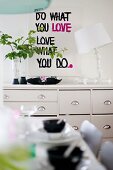 Table lamp with white lampshade and leafy twigs in vase on chest of drawers below motto on wall and behind set table