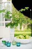 Vase mit Blätterzweig und Schale mit grünen Äpfeln, davor Windlichter auf Gartentisch