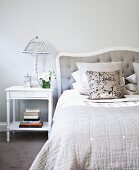 Table lamp on white bedside table next to French bed with pillows stacked against upholstered headboard in traditional bedroom painted pale grey