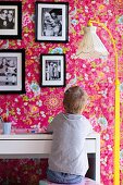 Kind am Schreibtisch, daneben Stehleuchte mit nostalgischem Stoffschirm und gelb lackiertem Gestell, Familienfotos an tapezierter Wand mit Blumenmuster auf pinkfarbenem Hintergrund