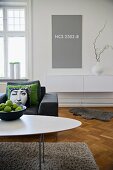 View across oval coffee table and armchair with stack of scatter cushions to white sideboard below grey panel with NCS colour code on wall