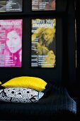 Detail of bed with yellow and black and white patterned scatter cushions on black throw and collection of posters on wall