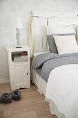 White wooden bedside table next to bed with tall headboard and grey and white bed linen
