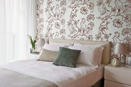 Elegantes, helles Schlafzimmer mit Vogelmotiv auf tapezierter Wand, davor Doppelbett mit Kopfpolster und Kissenstapel