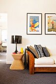 Framed retro posters above wicker sofa next to black table lamp on designer stool