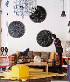 Yellow stool and wooden coffee table on animal-skin rug in front of sofa below collection of vintage clocks on wall