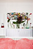 View across pink fur rug to white sideboard with roller doors and glass vases of tropical flowers below pin board on wall