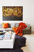 Low white coffee table on animal-skin rug, orange blanket and retro-style scatter cushions on sofa below modern painting on white wall