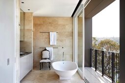 Free-standing bathtub next to open, sliding balcony door in modern bathroom with sand-coloured tiles on walls and floor