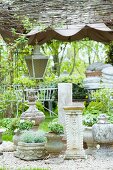 Stone pots and antique-style pillars for sale in a gravelled area of a garden with lanterns and white metal chairs in the background