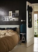 Bed with upholstered headboard below photos tacked on black-painted wall next to open interior door