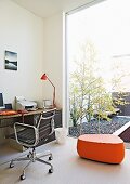 Bürostuhl-Klassiker mit schwarzer Lederbespannung und orangefarbenes Designer Möbel in Homeoffice; Glasfront zu Innenhof mit Birken in Kiesbeet