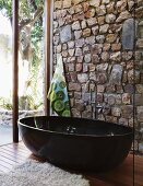 Designer Badezimmer mit schwarzer freistehender Wanne vor Natursteinwand und Glasfassade mit Blick auf Gartenterrasse