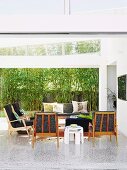 Sitzmöbel im 60er Jahrestil auf Terrazzoboden in hellem Wohnraum; Blick durch eine breite Glasschiebefront auf Bambushecke vor Sichtschutzzaun