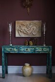 Elegant, silver candlesticks on vintage-style console table with drawers below antique low-relief on aubergine-coloured wall